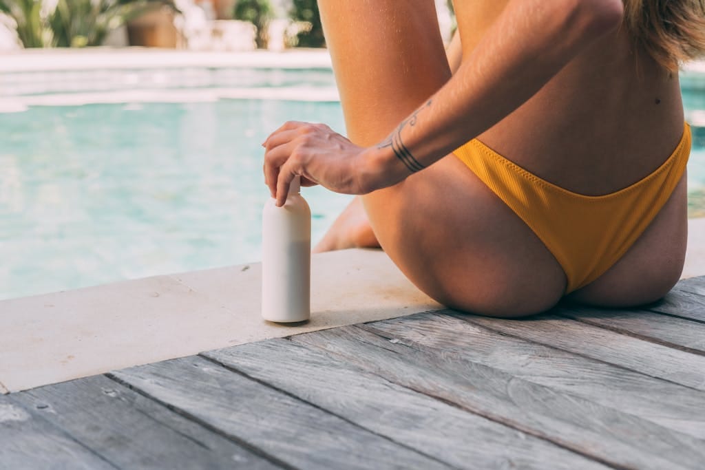 A woman sitting by the poolside, holding a sunscreen bottle in summer sunlight.