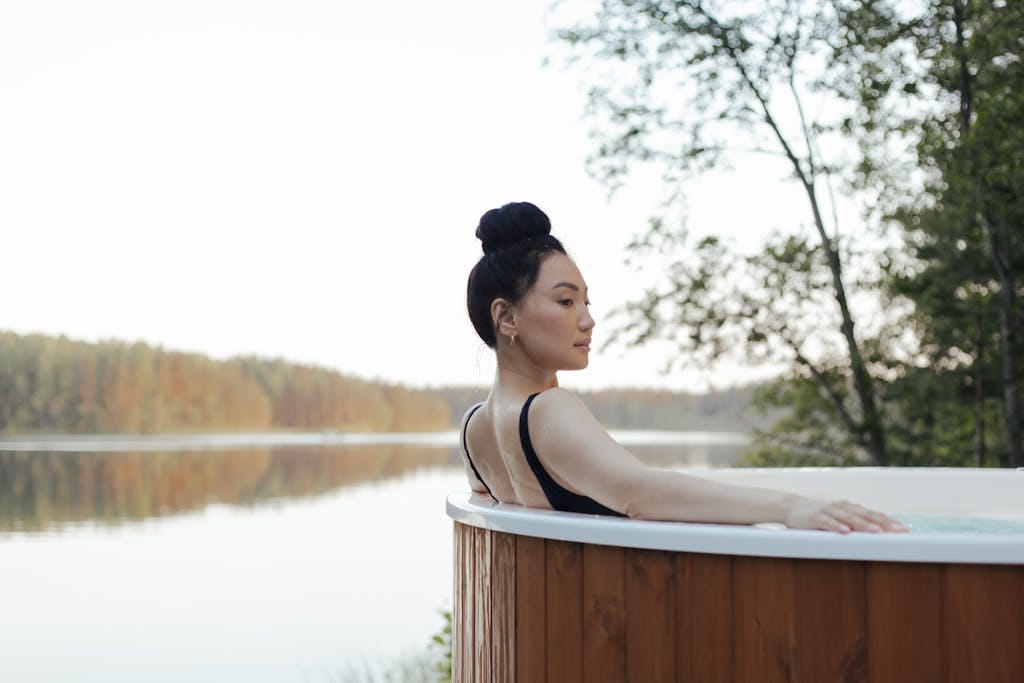 Asian woman enjoying a serene moment in a hot tub overlooking a tranquil lake.