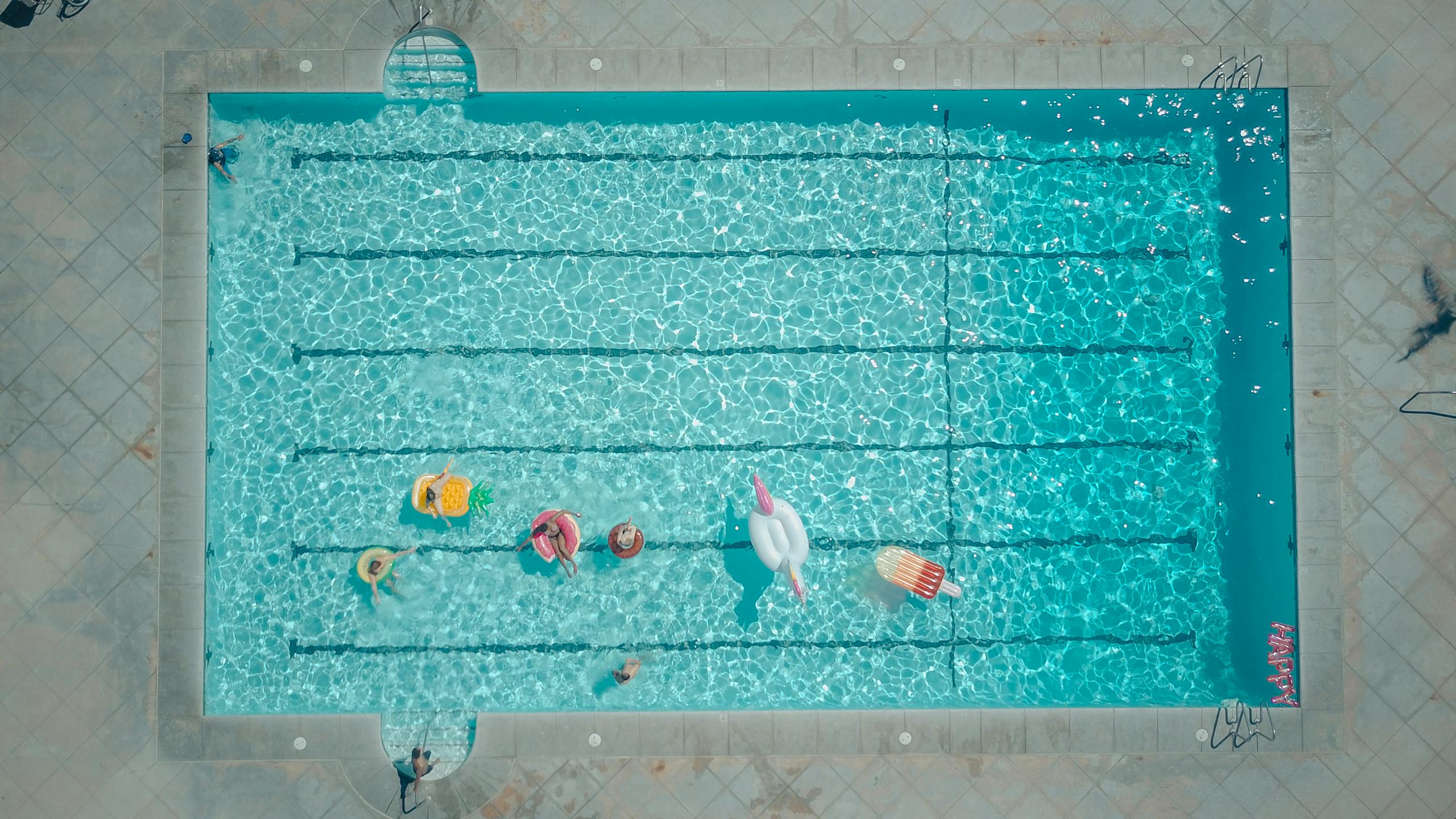Top-down view of a swimming pool with colorful floats and people enjoying a sunny pool party.